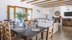 A dining area with a wooden table set for six, surrounded by wooden chairs. A white sofa and a television are visible in the background. - Casa de los Sueños (Javea / Xàbia)