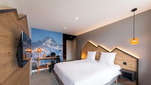 A modern hotel room with a large bed, a desk with a chair, a mounted TV, and a mountain mural on the wall.