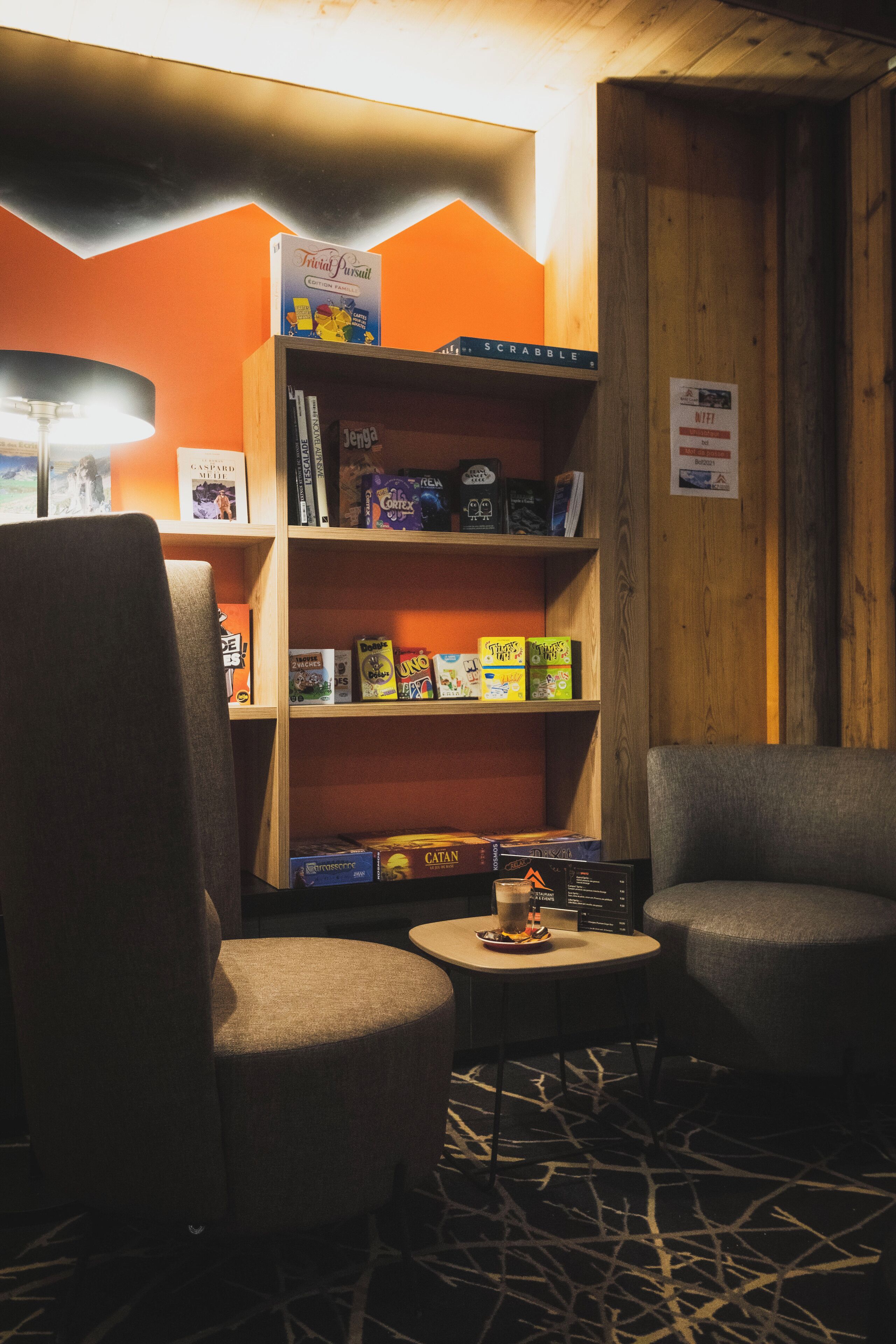 A cozy seating area with two armchairs and a small table, a bookshelf with various board games, and a lamp.