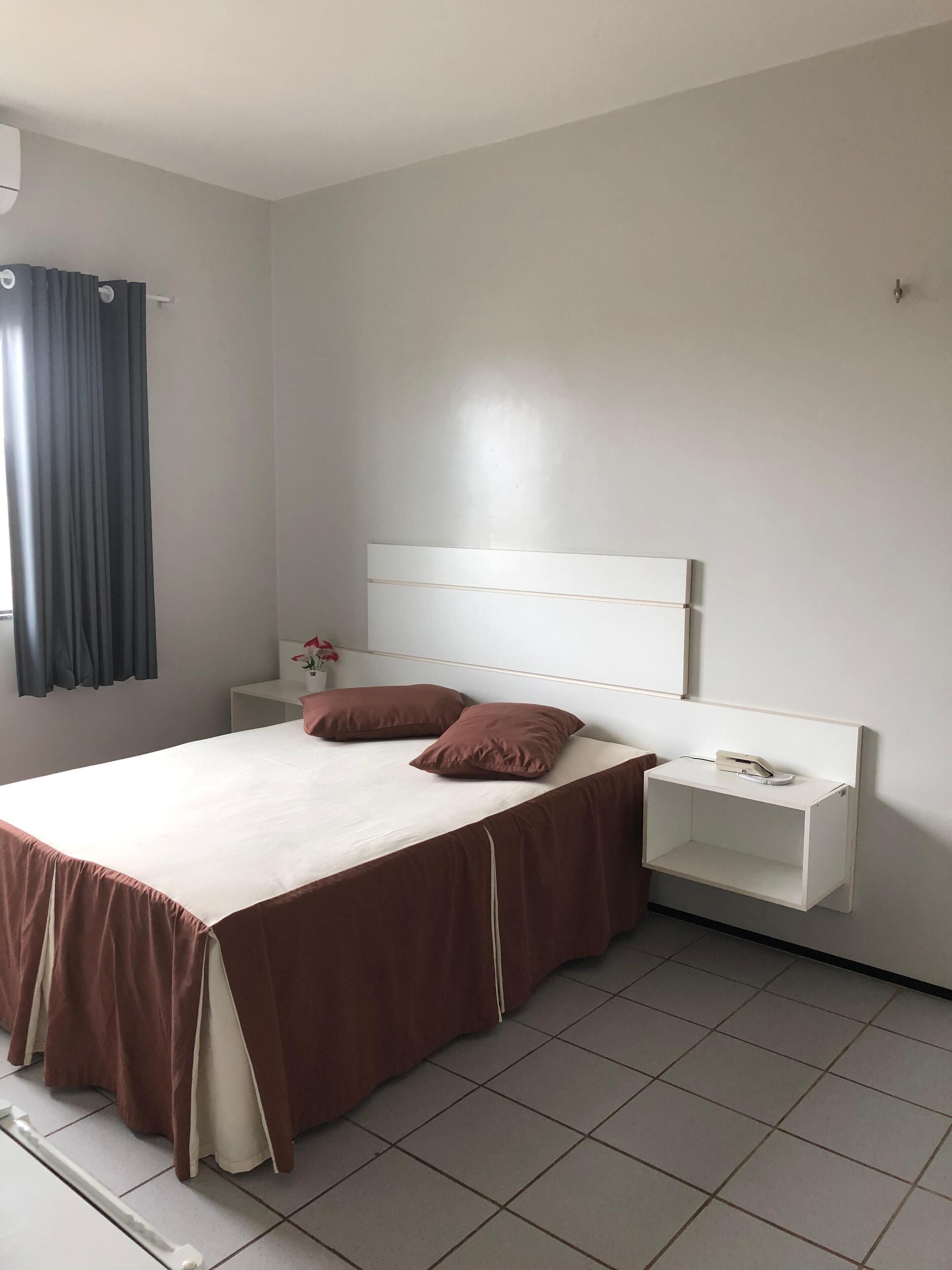 A bedroom with a bed, a nightstand, a wall-mounted shelf, and a window with curtains.