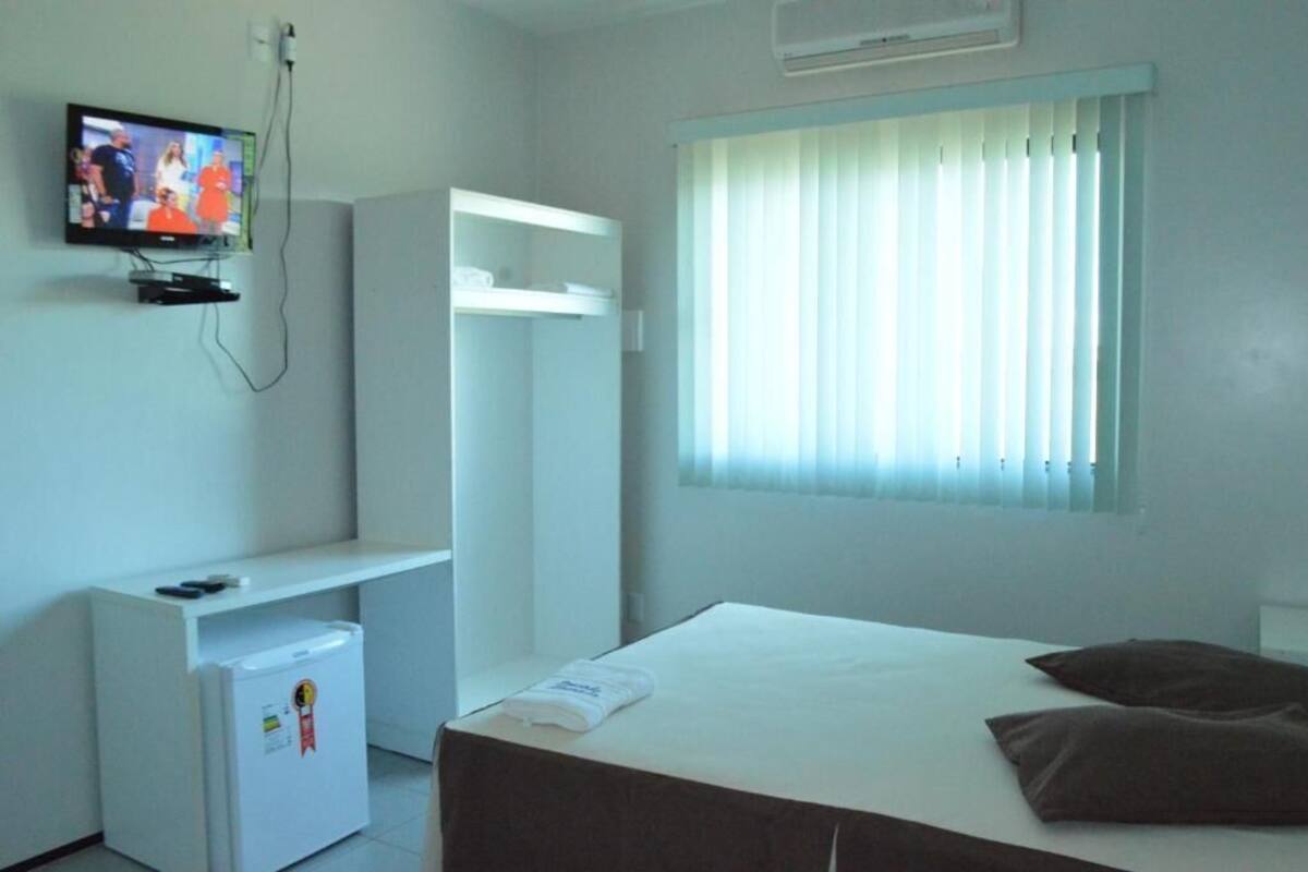 A hotel room with a bed, a TV mounted on the wall, a window with blinds, and a small refrigerator.