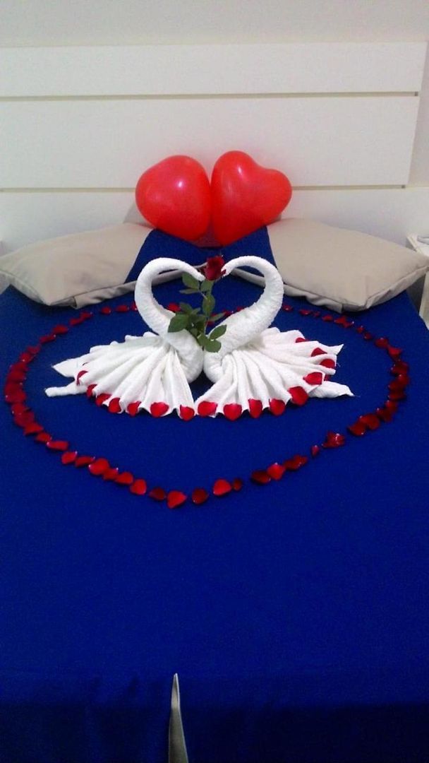 Cama com cobertura azul, almofadas brancas e arranjo decorativo de pétalas de rosa vermelhas e cisnes brancos.