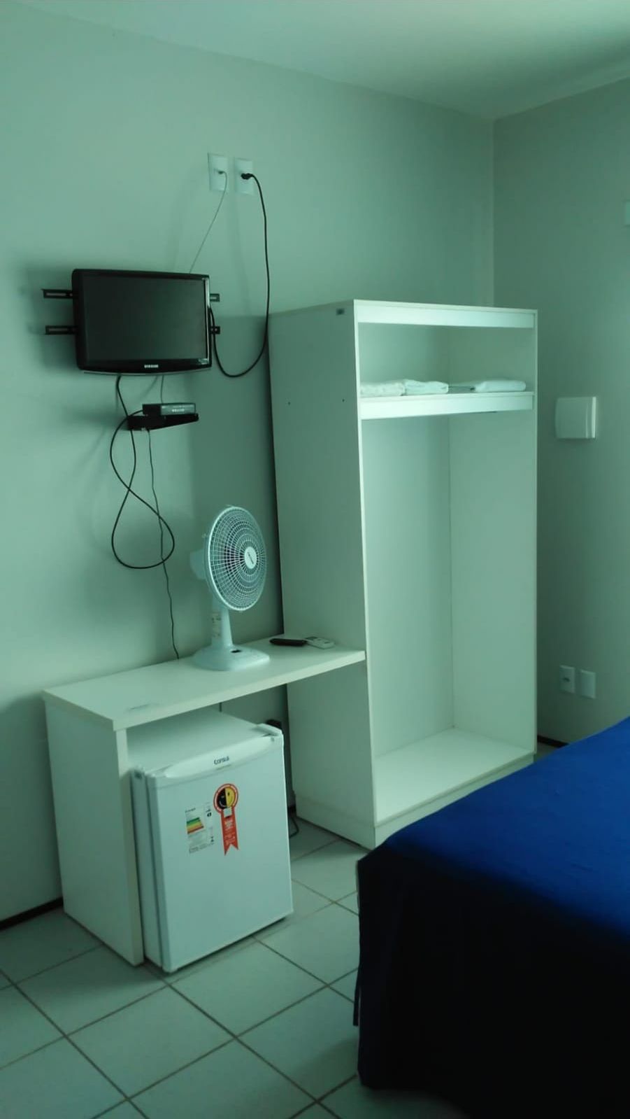 A small, sparsely furnished room with a bed, a desk with a computer and fan, and a white wardrobe.