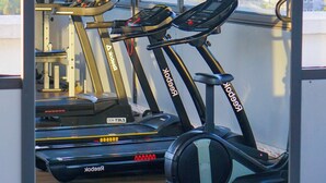Een sportschool met drie loopbanden en een hometrainer, grote ramen en uitzicht op een stadsbeeld.