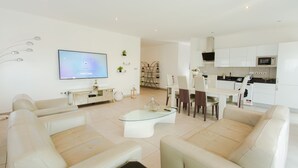 Een moderne woonkamer met een glazen salontafel, witte banken, een flatscreen televisie en een keuken met witte kasten en apparatuur.
