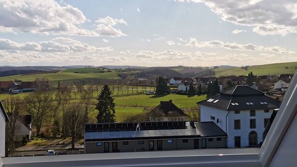 Vista da janela sobre paisagem rural com casas, campos verdes e céu parcialmente nublado.