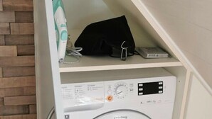 Máquina de lavar compacta e branca com painel de controle, situada numa pequena área de lavandaria fechada com chão de madeira e parede de tijolo.