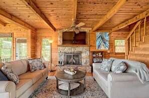 Cozy cabin interior with a central fireplace, comfortable seating area, and a wooden staircase leading to an upper level.