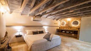 A bedroom with a large bed, wooden beams on the ceiling, a brick wall with a mirror, and a small nightstand with a lamp.