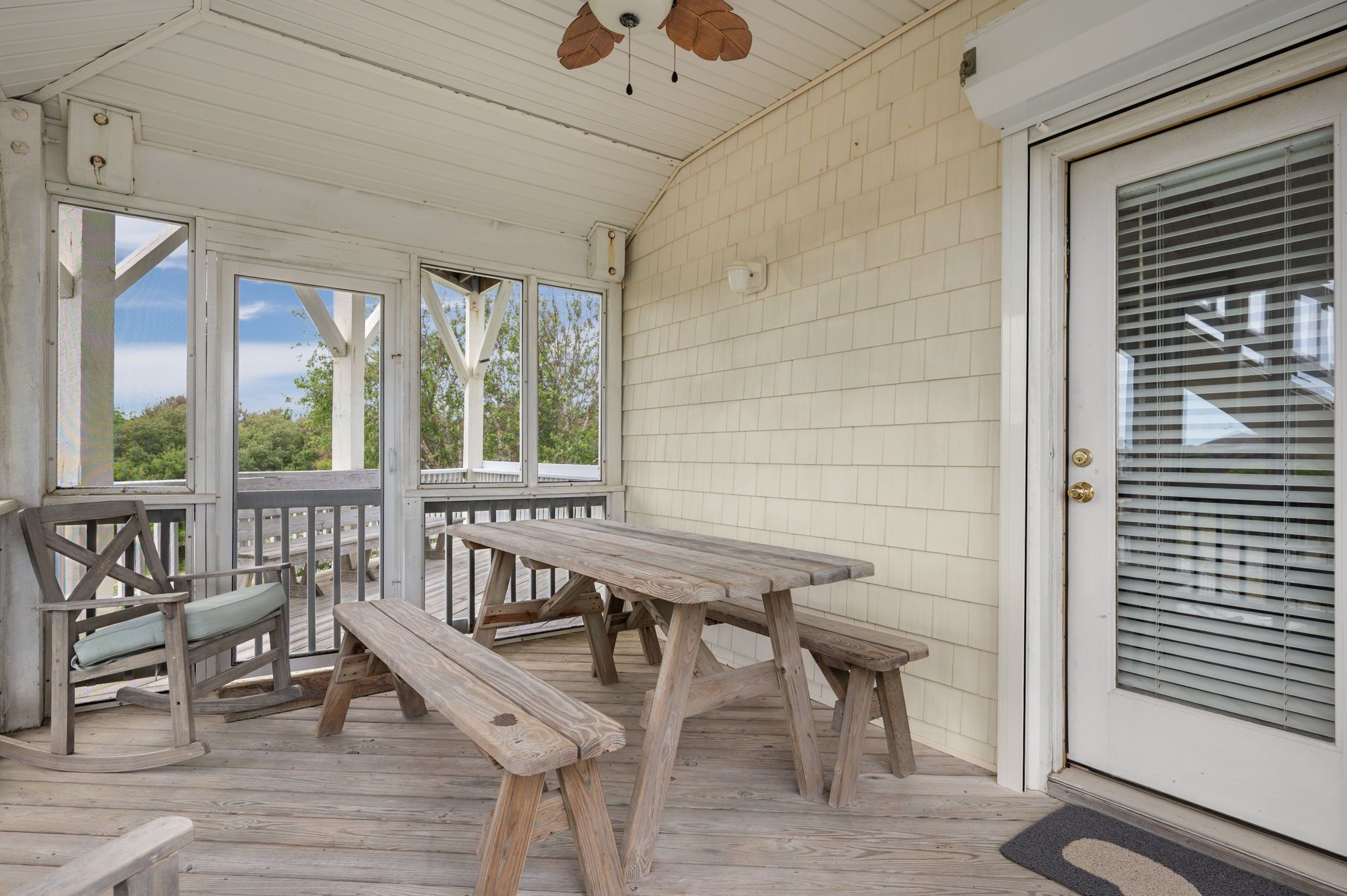 A covered outdoor area with wooden picnic tables and benches, a white door with a window, and a ceiling fan.
