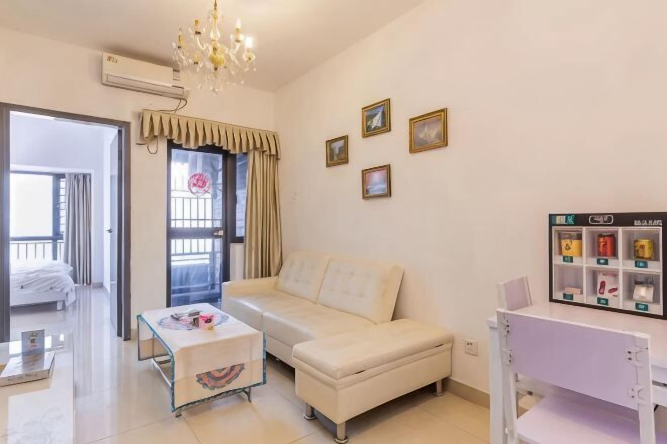 Una habitación con un sofá blanco, una mesita, una silla blanca, una araña, una ventana con cortinas y un estante con marcos.