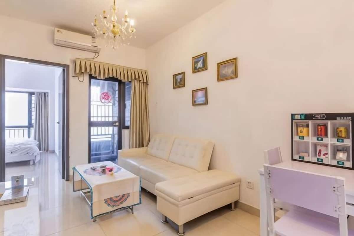 Una habitación con un sofá blanco, una mesita, una silla blanca, una araña, una ventana con cortinas y un estante con marcos.