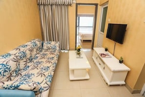 Una habitación con un sofá floral, una mesa de centro blanca con un jarrón, una mesita auxiliar blanca, un televisor montado en la pared y una puerta que da a un balcón.