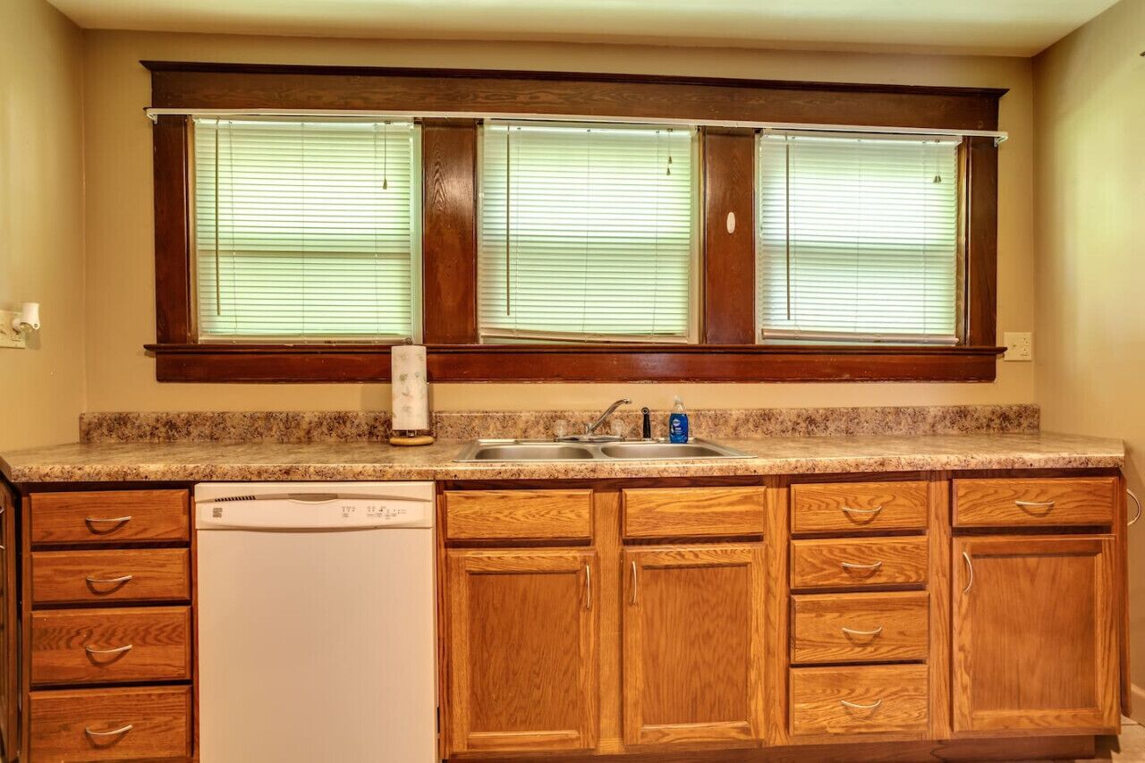 A kitchen with wooden cabinets, a built-in dishwasher, and a window with blinds.