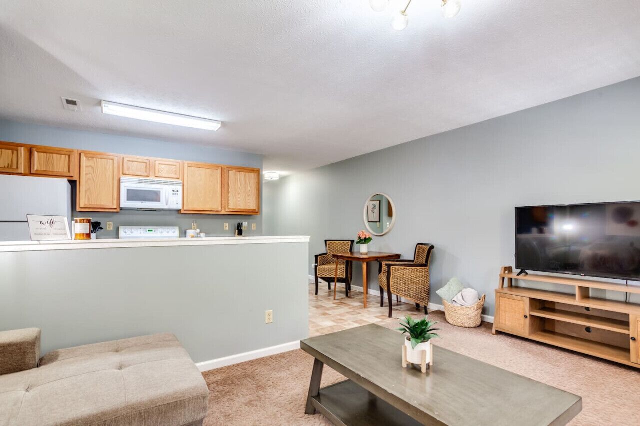 A modern living room with a flat-screen TV, a dining area with wicker chairs, and a kitchen with wooden cabinets and a microwave.