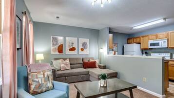 A living room with a sectional sofa, a coffee table, and a kitchen area with wooden cabinets and a microwave.