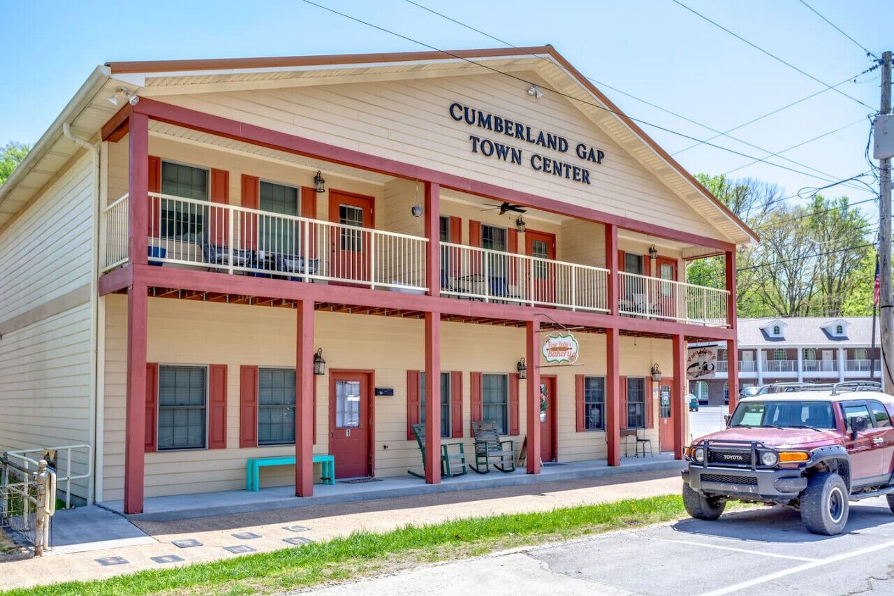 Um edifício de dois andares com varanda, mesas ao ar livre e um veículo estacionado, identificado com a placa 'Cumberland Gap Town Center'.