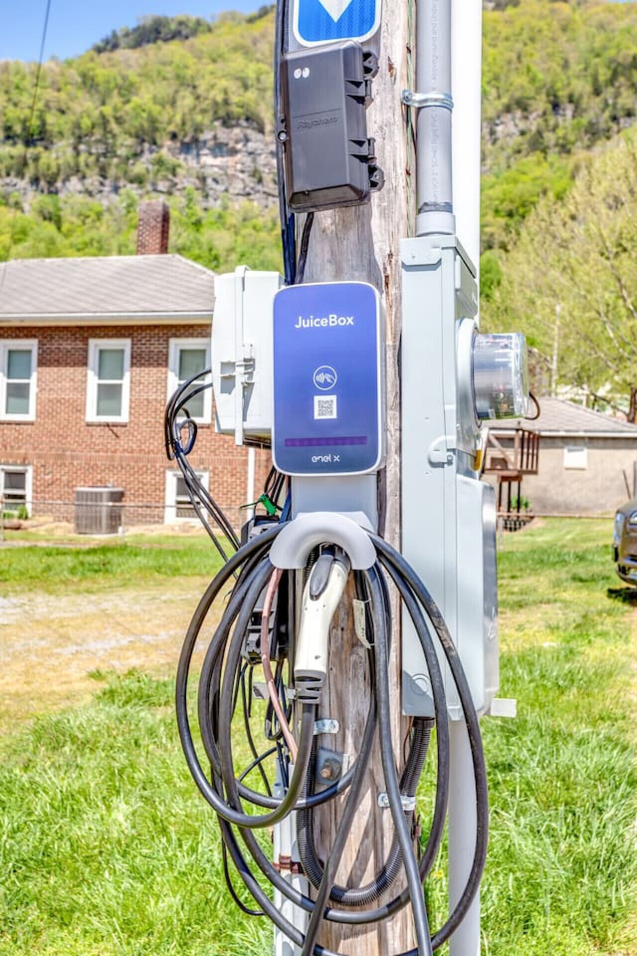 A JuiceBox charging station with multiple cables connected to it, set against a backdrop of a brick building and a grassy area.