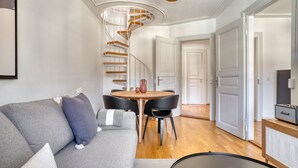 A modern living room with a grey sofa, a round wooden table, and black chairs. A spiral staircase with wooden steps leads to an upper floor. - Charming Breite 3 Room w/ Washer & Storage, nr Market, by Blueground (Basel)