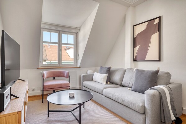 A modern living room with a grey sofa, a pink chair, a coffee table, and a TV. - Charming Breite 3 Room w/ Washer & Storage, nr Market, by Blueground (Basel)