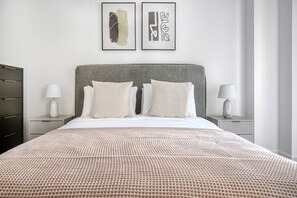 Un lit bien fait, recouvert d’une couvre-lit à motifs, avec deux oreillers et une tête de lit grise. Deux tables de chevet, sur lesquelles sont posées des lampes, se trouvent de part et d’autre du lit, et des œuvres d’art encadrées sont accrochées au-dessus.