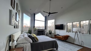Un dormitorio con cama, televisor, chimenea y ventanas amplias.