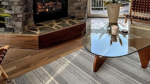 Una sala de estar con chimenea de piedra, una mesa de centro de vidrio y vistas a árboles a través de amplios ventanales.