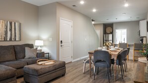 A modern living room with a gray sectional sofa, a wooden dining table with chairs, and a kitchen area in the background.