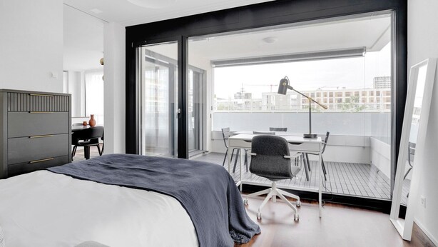 A modern bedroom with a large bed, a desk, and a chair. It has a balcony with a view of the city. - Luxe Opfikon 4.5 Room w/ Storage & W/D, 2 mins to Market, by Blueground (Zurich)