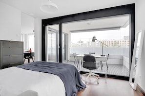 A modern bedroom with a large bed, a desk, and a chair. It has a balcony with a view of the city.