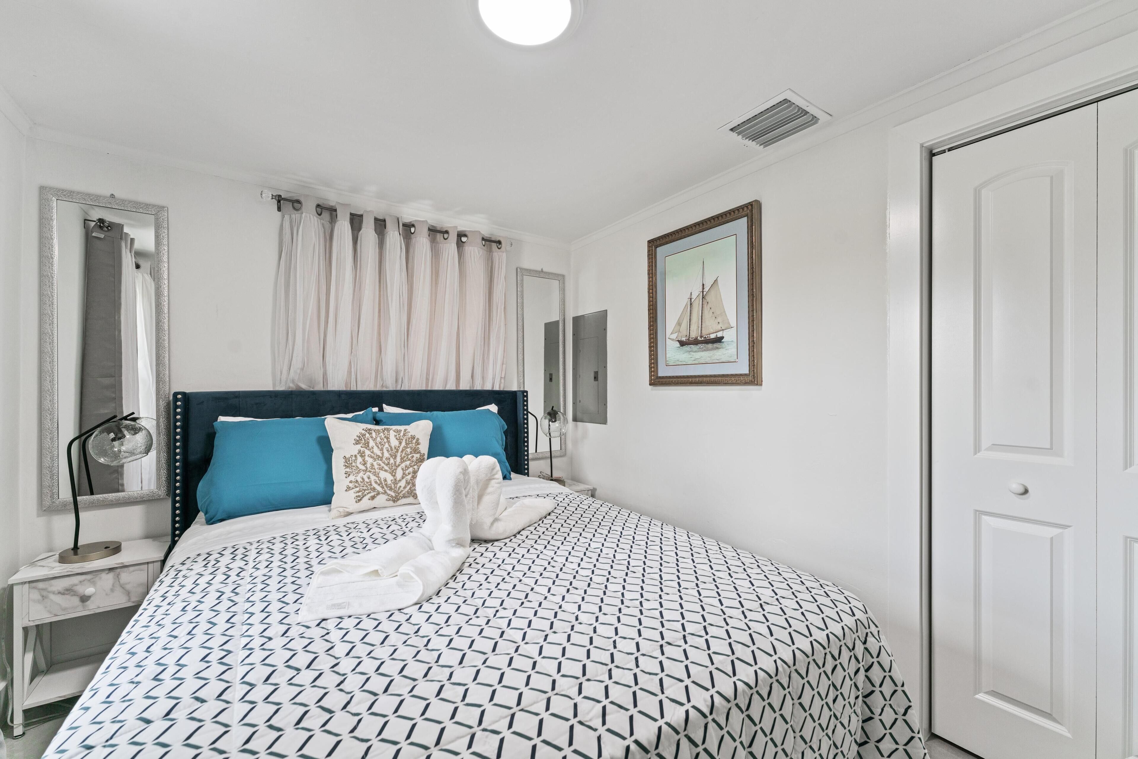 A neatly arranged bedroom with a patterned bedspread, a headboard, a nightstand, a mirror, a framed picture on the wall, and a closed door.