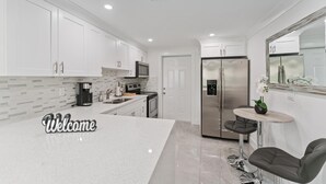 Una cocina moderna con armarios blancos, un refrigerador de acero inoxidable, una cafetera y un cartel de bienvenida.