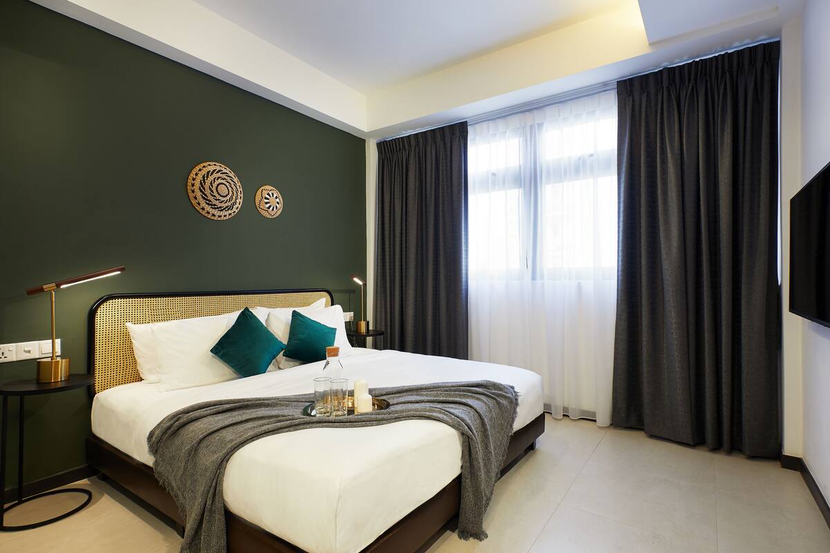 A neatly made bed with a grey throw blanket, two pillows, and a tray with glasses and a candle. There is a bedside table with a lamp and a small decorative item. The room has dark curtains and a large window with sheer white curtains.