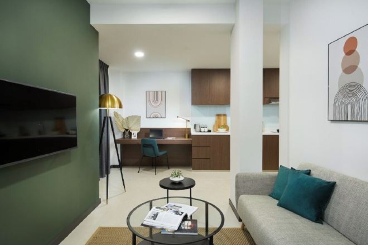 A modern living room with a sofa, a coffee table, and a TV. There is a kitchen area in the background with a desk and cabinets.