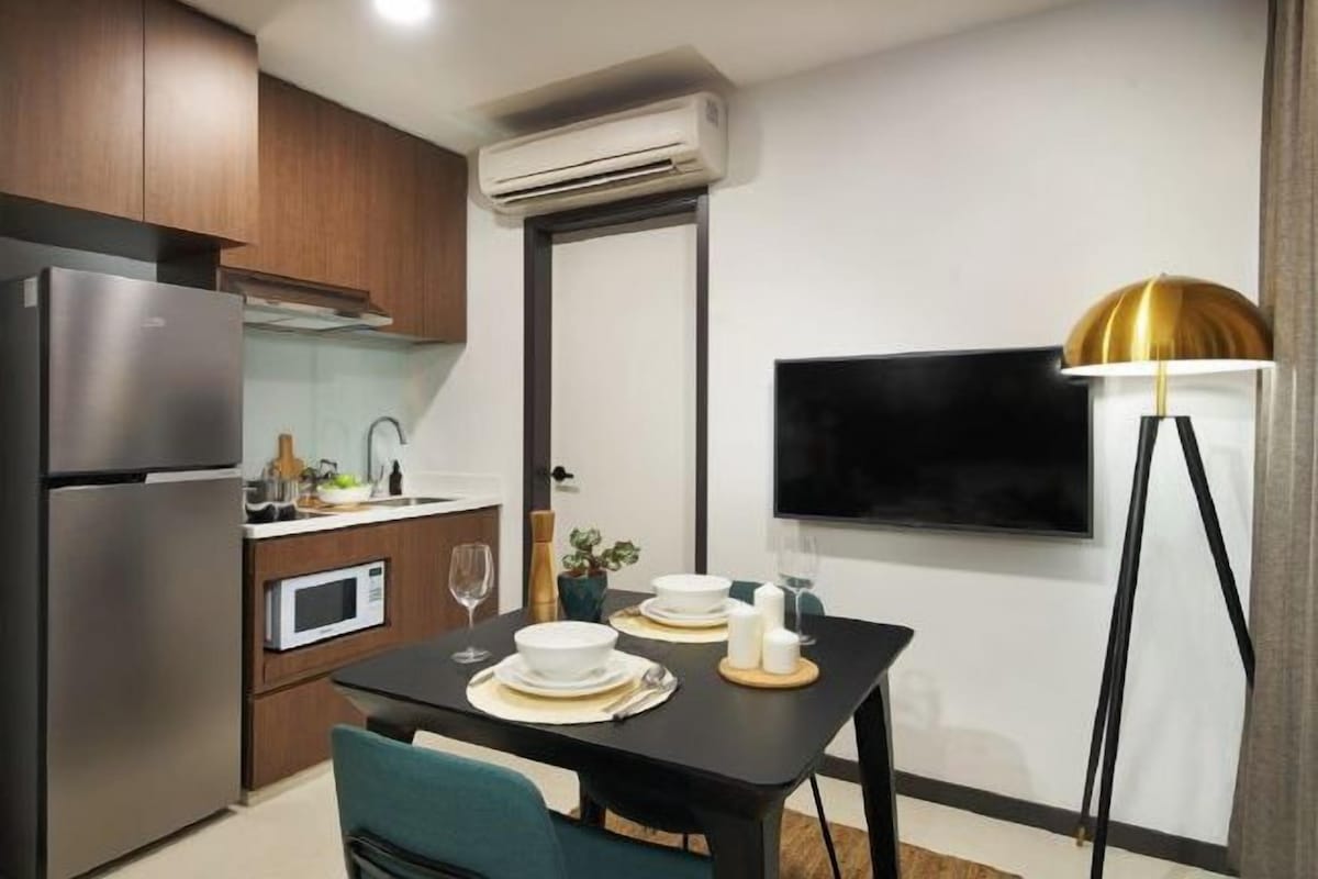 A compact kitchen with a refrigerator, microwave, and a dining area set with a table, chairs, and a TV mounted on the wall.