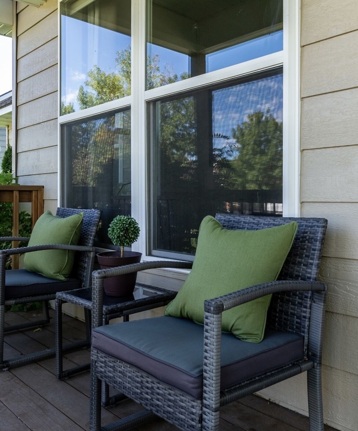 A porch with wicker chairs and cushions, a small potted plant, and a window with a view of trees.