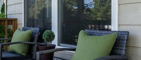 A porch with wicker chairs and cushions, a small potted plant, and a window with a view of trees.