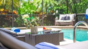 Un área junto a la piscina con una tumbona, una mesa pequeña con un frutero y una planta en maceta.