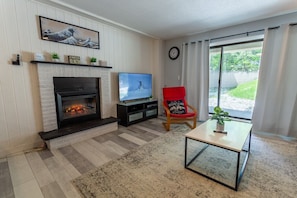 Un salón con chimenea, televisor, una butaca roja y vistas al verdor exterior.