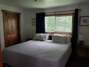 A bedroom with a large bed, a wooden door, a window with curtains, and a bedside table with a lamp.