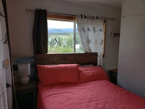 A bedroom with a wooden headboard, a bed with pink bedding, a window with curtains, and a view of a forested area.