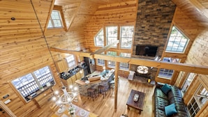 Une cabane spacieuse avec un plafond haut, des murs en bois, une cheminée en pierre et de grandes fenêtres.