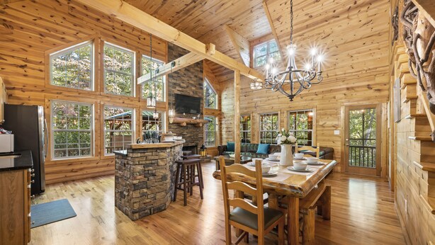 Une vaste cabane en rondins dotée d’une cheminée en pierre, d’une table à manger en bois et de grandes fenêtres offrant une vue sur la forêt environnante.