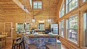 Une cuisine rustique, située dans une cabane en bois, avec un îlot central en pierre, des tabourets de bar en bois et de grandes fen êtres.