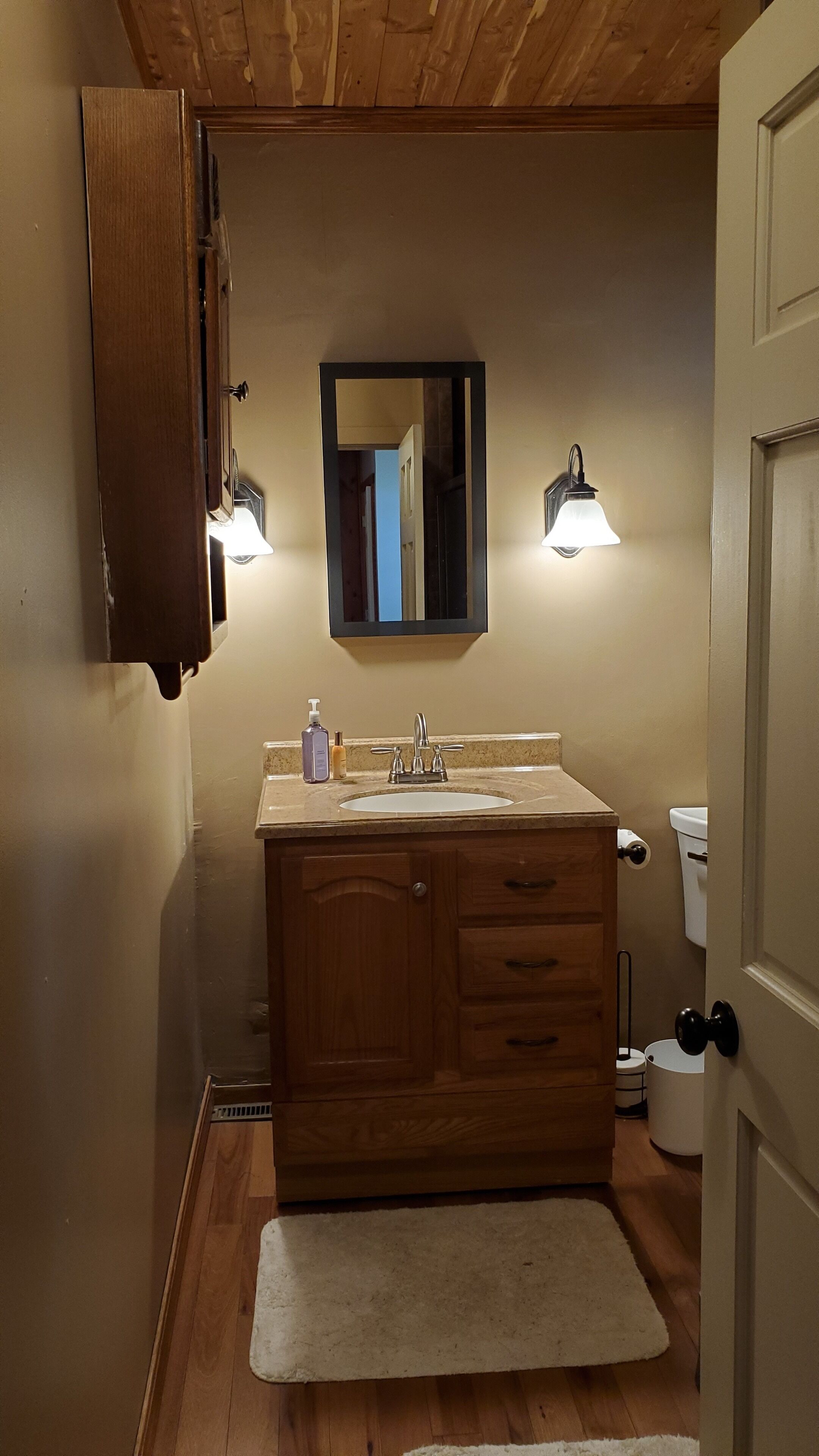 Une salle de bain avec un meuble-lavabo en bois, un évier, un miroir, deux appliques murales, des toilettes et un tapis blanc.