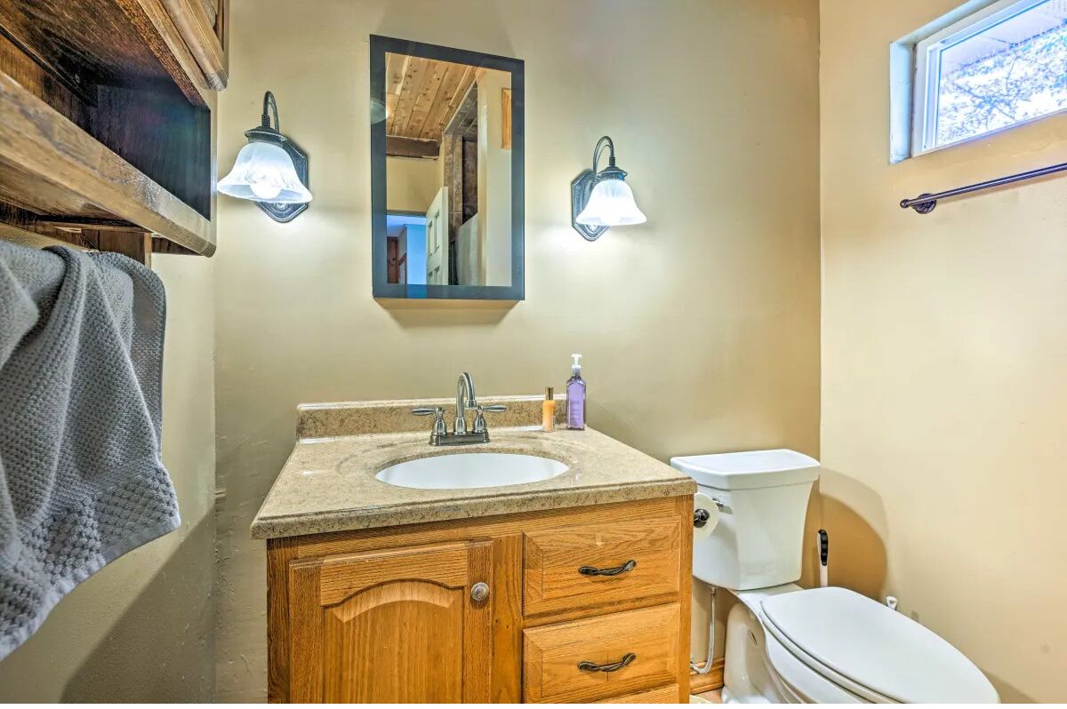 Baño con tocador de madera, lavamanos y retrete. Hay dos luces en la pared y un espejo sobre el lavamanos.