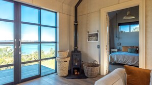 A room with a large sliding glass door leading to a balcony, a wood-burning stove, a bed with a view, and a cozy seating area. - Loft & Workroom, Woodside Bay | Be My Guest (Auckland)