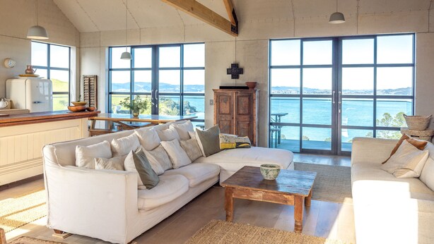 A spacious living area with a large white sofa, a wooden coffee table, and a view of the sea through large windows. - Loft & Workroom, Woodside Bay | Be My Guest (Auckland)