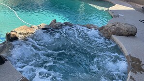 Ofurô com cascata, cercado por pedras, em área de piscina com deck revestido de azulejos.