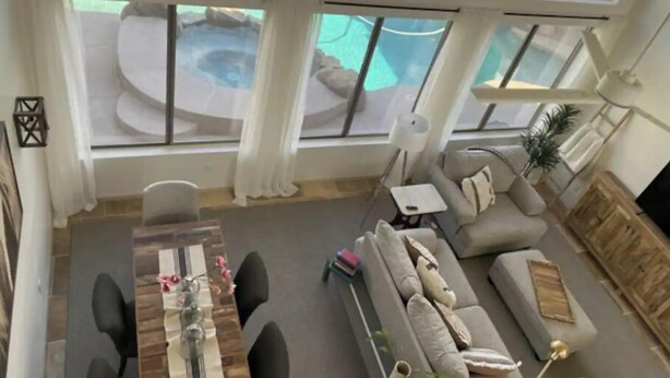 Ampla sala de estar com uma grande janela com vista para uma piscina, uma mesa de jantar com cadeiras e um sofá com almofadas.